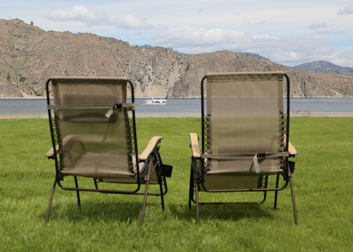 Two camp chairs on grass facing the lake and mountains, with a houseboat in the distance.
