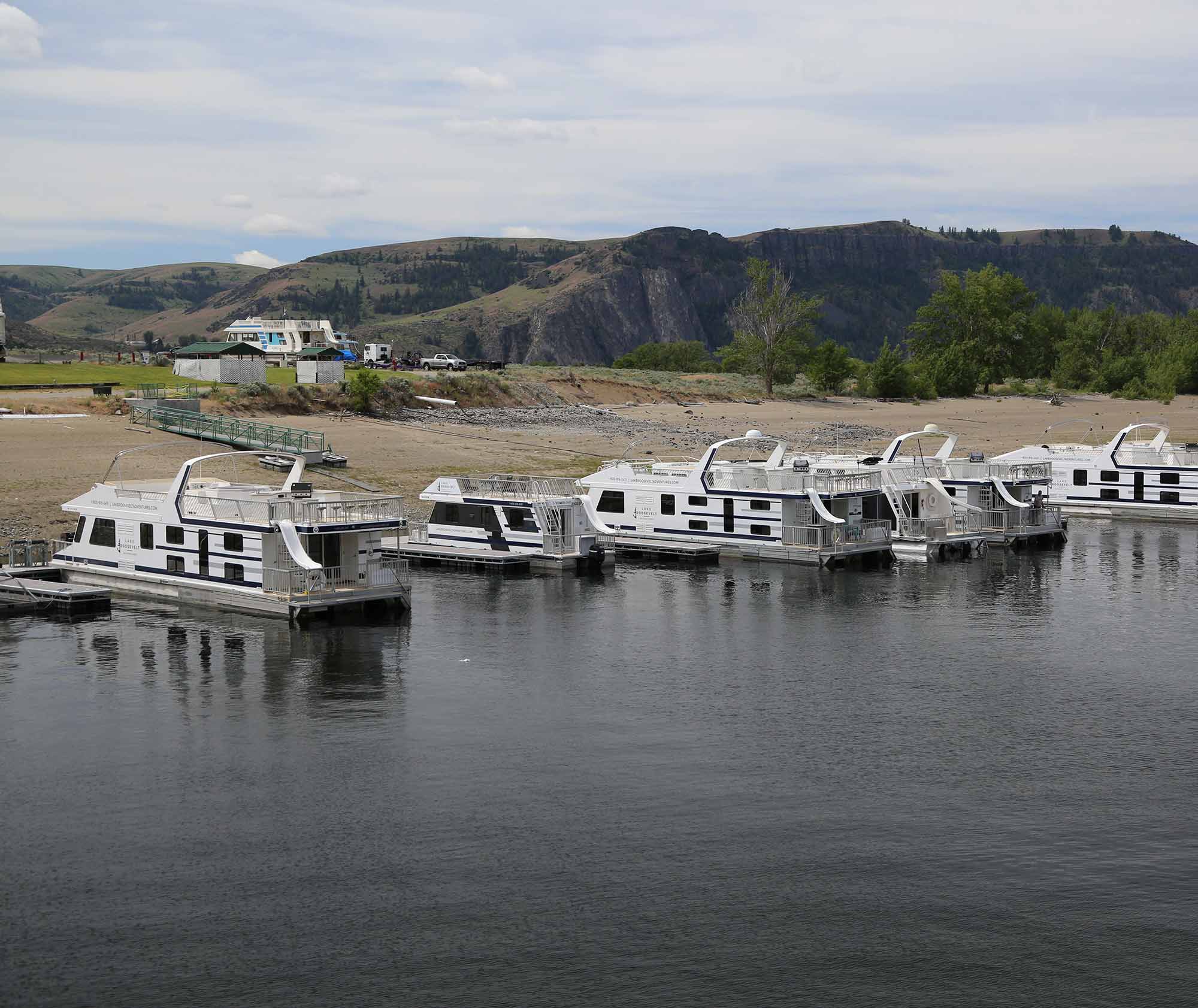 Several houseboats docked along a sandy shoreline with hills behind.